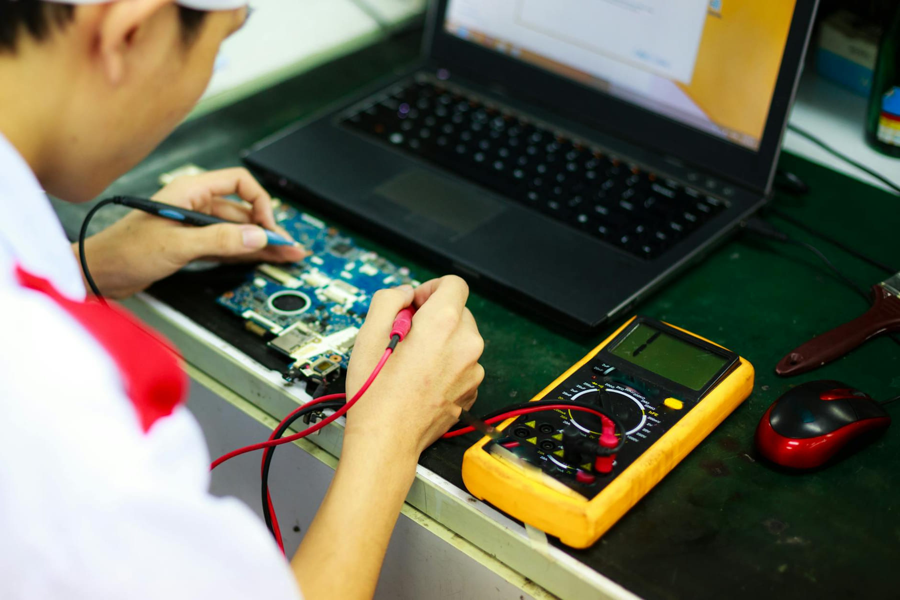 Technician working on a laptop with multimeter in a professional workspace. on The Tech Shelf